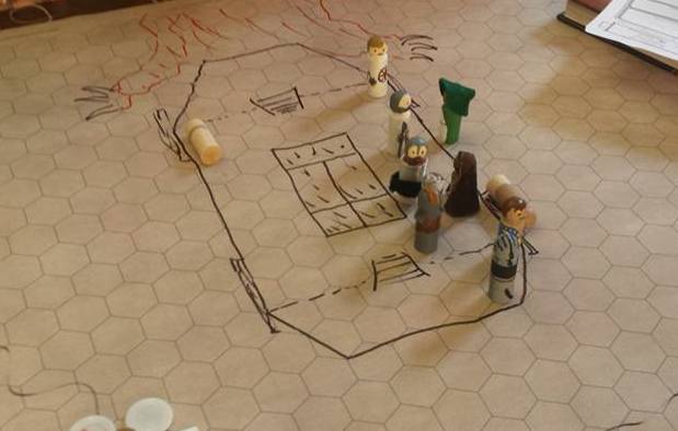 a gaming grid has the marker outline of a cart being pulled by a lizard. there are many miniatures made of clothespins on it in a battle for their lives